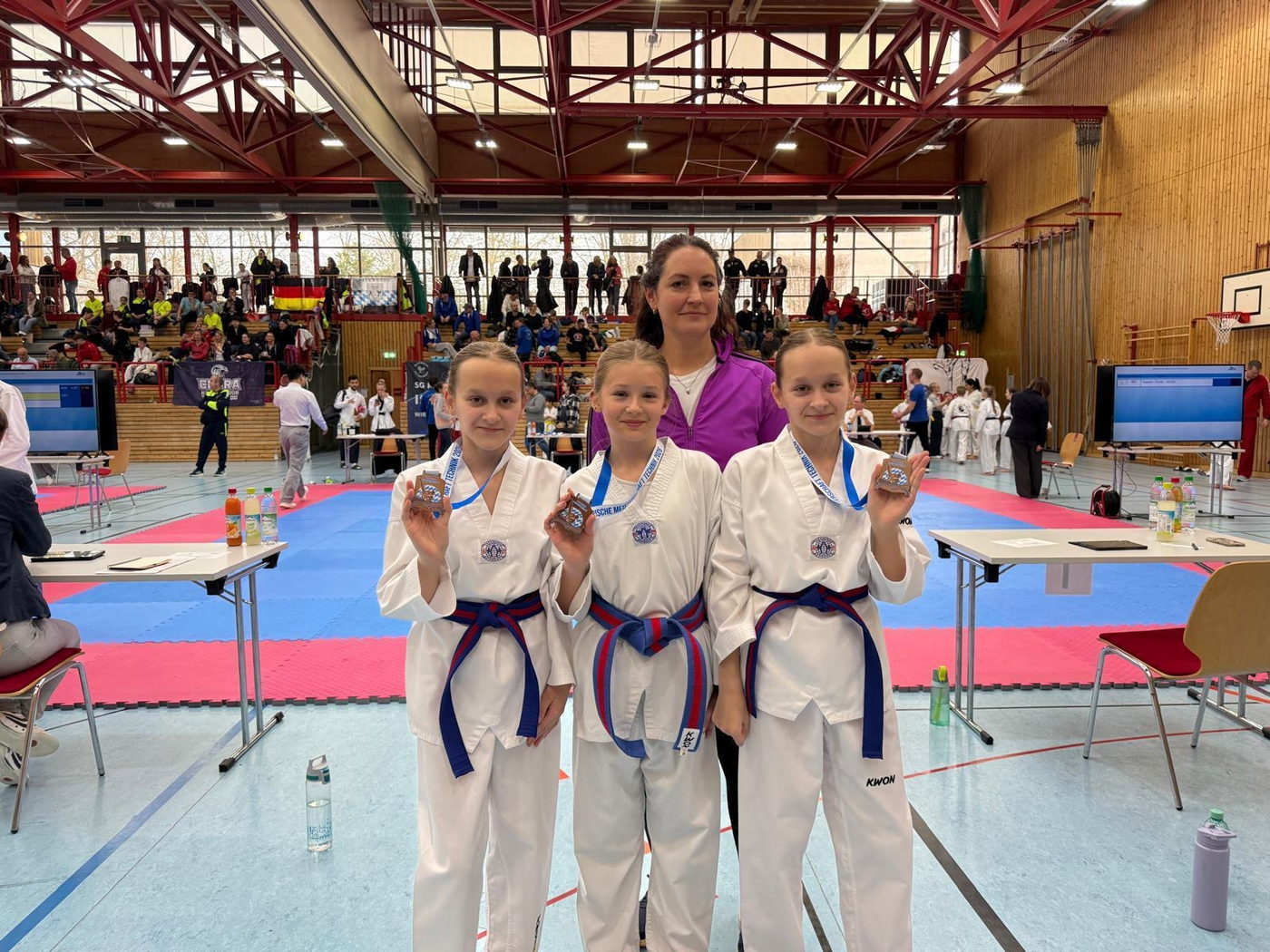 Drei junge Taekwondo-Sportlerinnen mit Medaillen und ihrer Trainerin beim Turnier